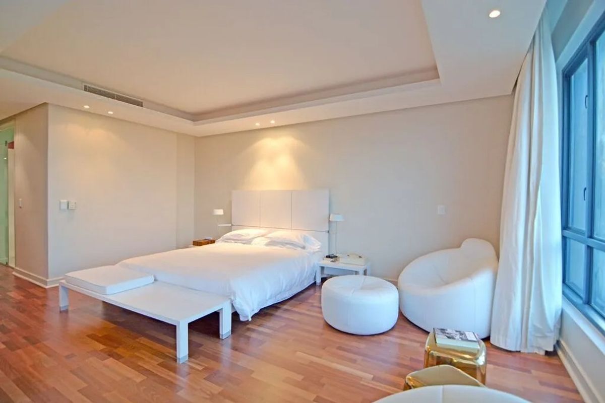 A spacious, well-lit bedroom featuring polished hardwood flooring, recessed ceiling lighting, and large windows with floor-to-ceiling curtains.