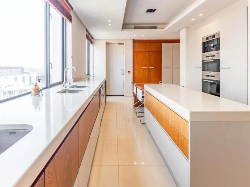 Modern penthouse kitchen featuring sleek white countertops, integrated cabinetry, built-in oven and microwave, and large windows offering expansive city views.