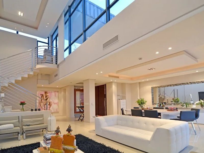 Spacious open-plan living and dining area featuring double-volume ceilings, floor-to-ceiling windows, recessed lighting, and a modern staircase with stainless steel railings.