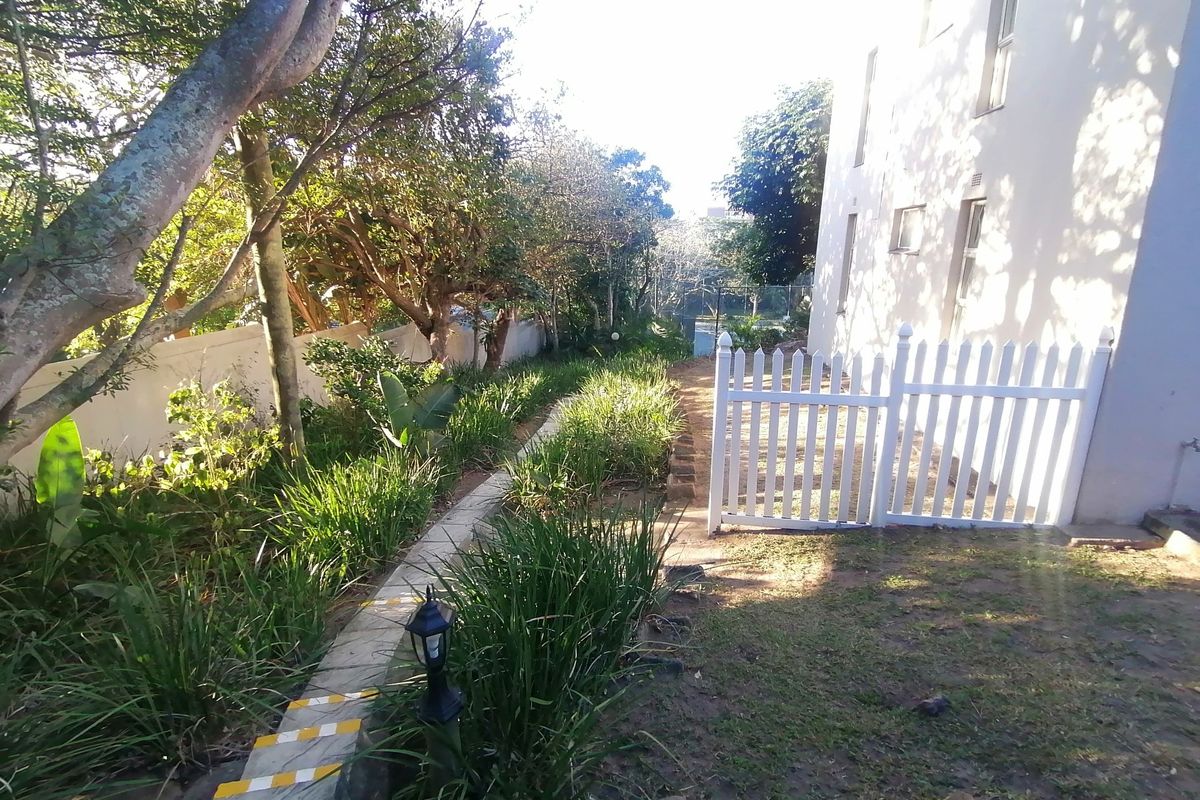 A side garden area featuring a paved walkway, established trees, and a white picket fence bordering the apartment building exterior.