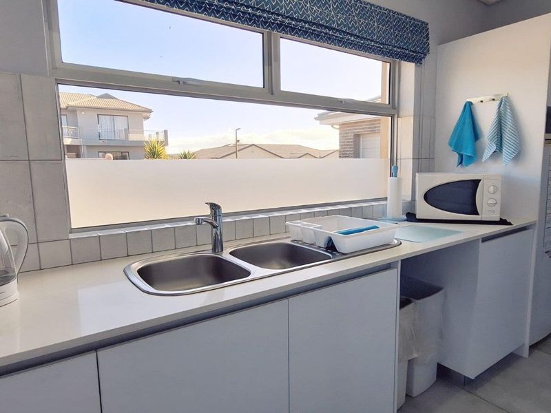 A functional kitchen scullery area featuring a double stainless steel sink, white cabinetry, tiled splashback, and a large window providing natural light.
