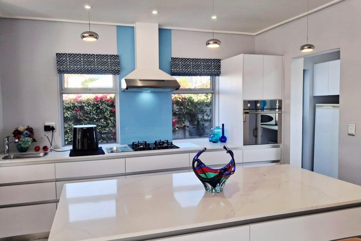 Modern kitchen featuring a central island with stone countertop, built-in oven, gas hob, extractor fan, and sleek cabinetry with a blue glass splashback.