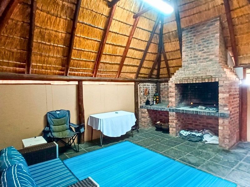 Covered outdoor entertainment area featuring a traditional thatched roof, exposed timber beams, and a built-in brick braai fireplace with stone tile flooring.