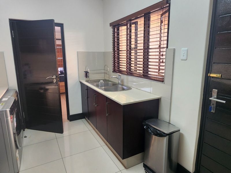 A functional scullery featuring dark wood cabinetry, stone countertops, a stainless steel double sink, and tiled flooring with direct access to adjacent rooms.