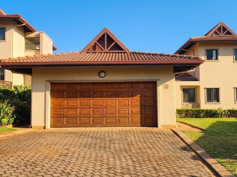 Front view of a townhouse featuring a double garage, paved driveway, and well-maintained garden with timber fencing.