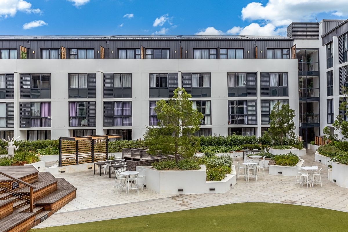 A well-maintained communal outdoor terrace featuring paved areas, raised garden beds, a built-in brick braai, and timber decking set against a modern apartment building.