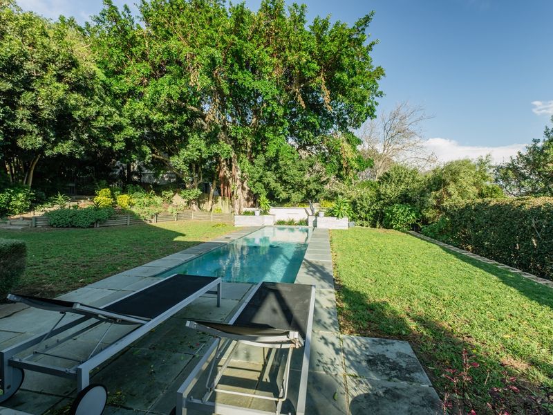 A rectangular swimming pool set within a lush, landscaped garden featuring stone paving and mature trees.