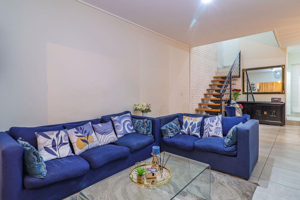 A bright living room featuring tiled flooring, a feature brick wall, and an open staircase with wooden treads and a metal railing.