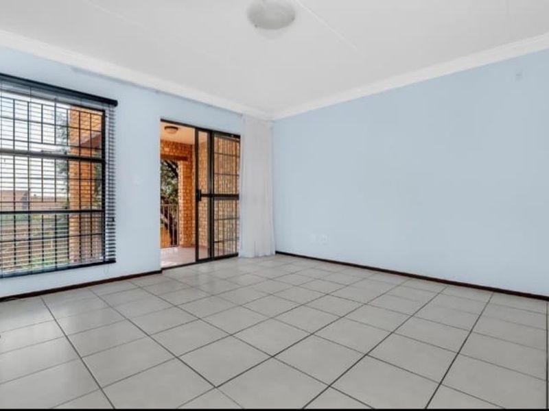 A bright, empty living room featuring neutral floor tiles, large windows with security bars, and sliding glass doors leading to an outdoor patio area.