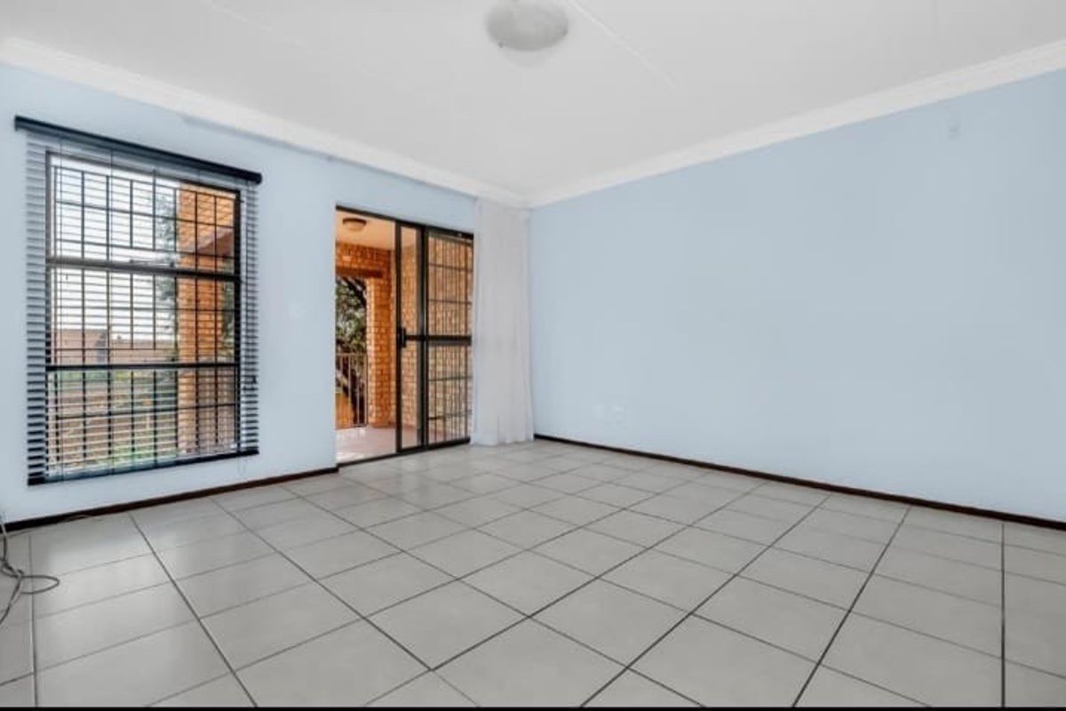 A bright, empty living room featuring neutral floor tiles, large windows with security bars, and sliding glass doors leading to an outdoor patio area.
