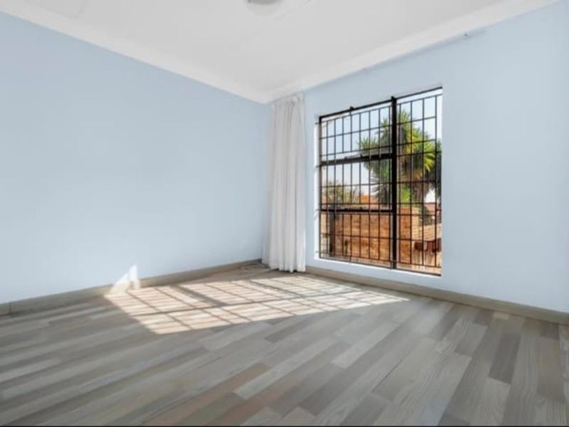 A bright, empty bedroom featuring light-toned wood-look flooring, pale blue walls, and a window secured with black metal burglar bars.