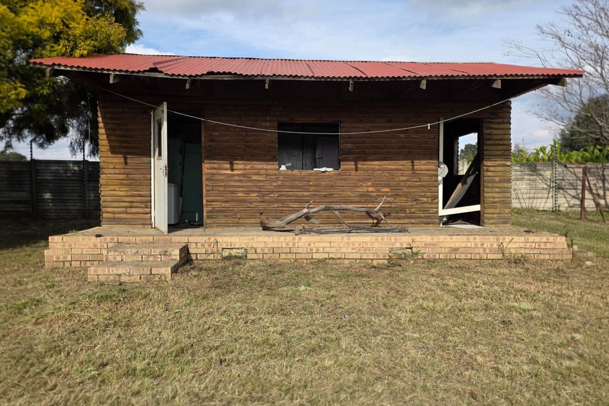 A rustic timber cabin structure featuring a corrugated metal roof and brick foundation, set within a large open garden area.