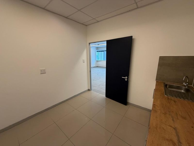 A compact office kitchenette area featuring tiled flooring, a sink with a wooden countertop, and a dark interior door leading to an adjacent office space.