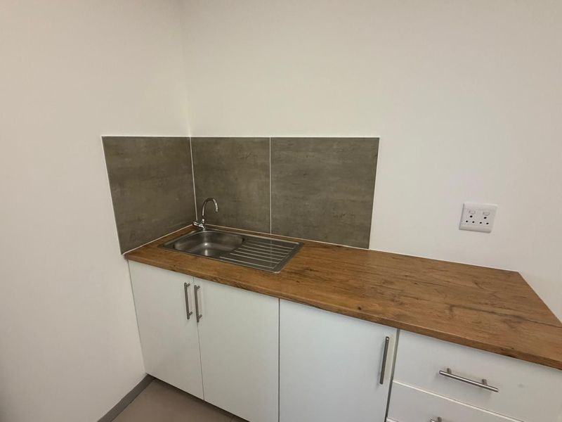 A compact office kitchenette featuring white cabinetry, a wood-look countertop, a stainless steel sink, and modern grey tiled splashback.