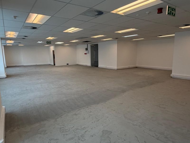 A spacious, open-plan office area featuring a suspended acoustic ceiling with integrated lighting, fire safety signage, and an unfinished concrete floor.