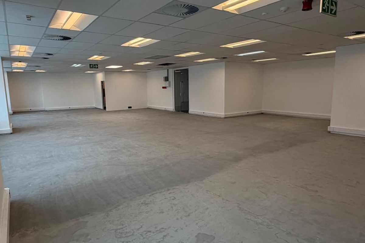A spacious, open-plan office area featuring a suspended acoustic ceiling with integrated lighting, fire safety signage, and an unfinished concrete floor.