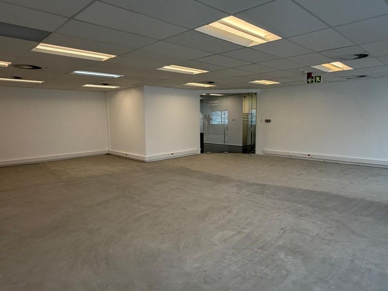 An open-plan office space featuring a suspended acoustic ceiling with recessed lighting, concrete flooring, and a glass entrance door.