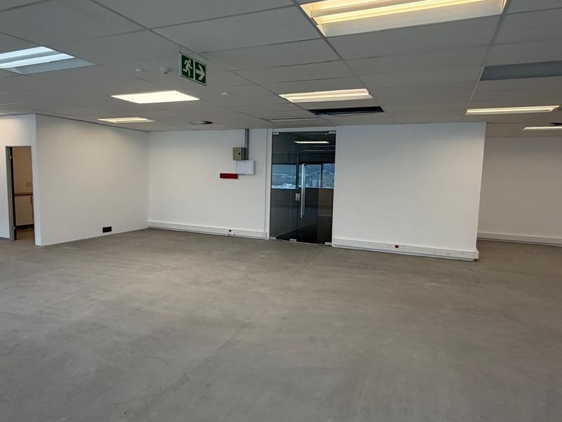 An open-plan office space featuring a polished concrete floor, suspended ceiling grid with recessed lighting, and a glass-paneled entrance door.