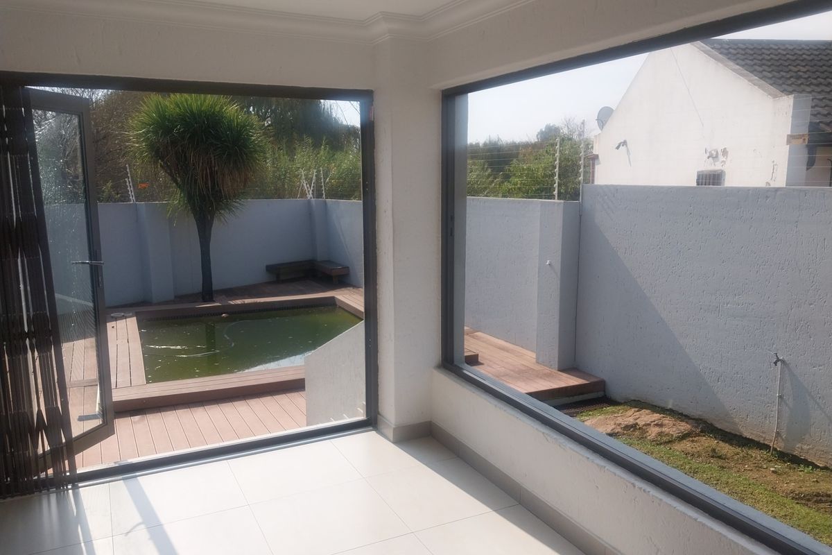 Bright interior living space with large glass doors opening onto a wooden deck, swimming pool, and private garden area.