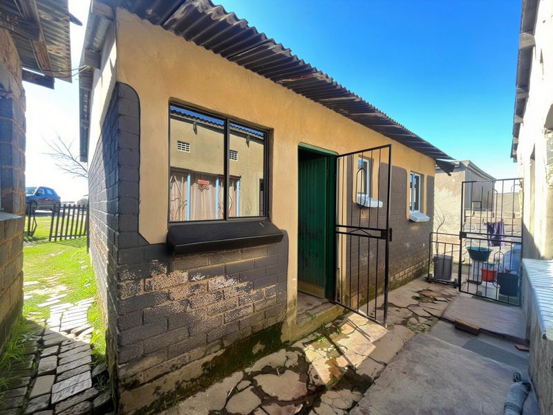 Rear exterior view of a small residential structure featuring concrete block walls, corrugated iron roofing, and security gate access.