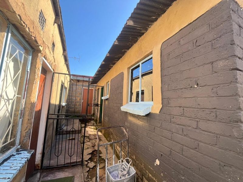 A narrow exterior side passage between two structures featuring brick walls, security gates, and corrugated iron roofing with visible signs of wear and maintenance needs.