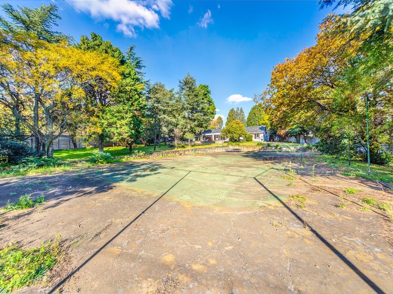 A large, weathered tennis court set within a spacious, tree-lined garden area, requiring significant resurfacing and maintenance to restore its functionality.