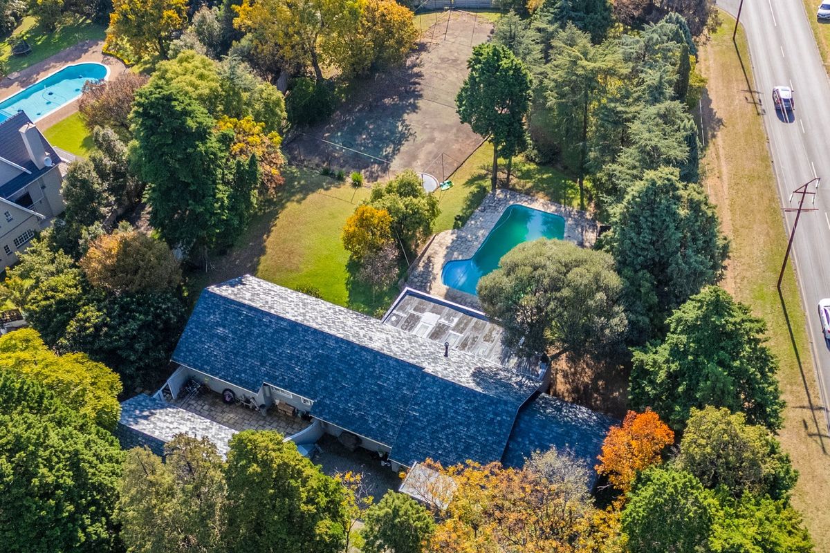 Aerial view of a large residential property featuring a swimming pool, expansive garden, and mature trees, situated near a main road.