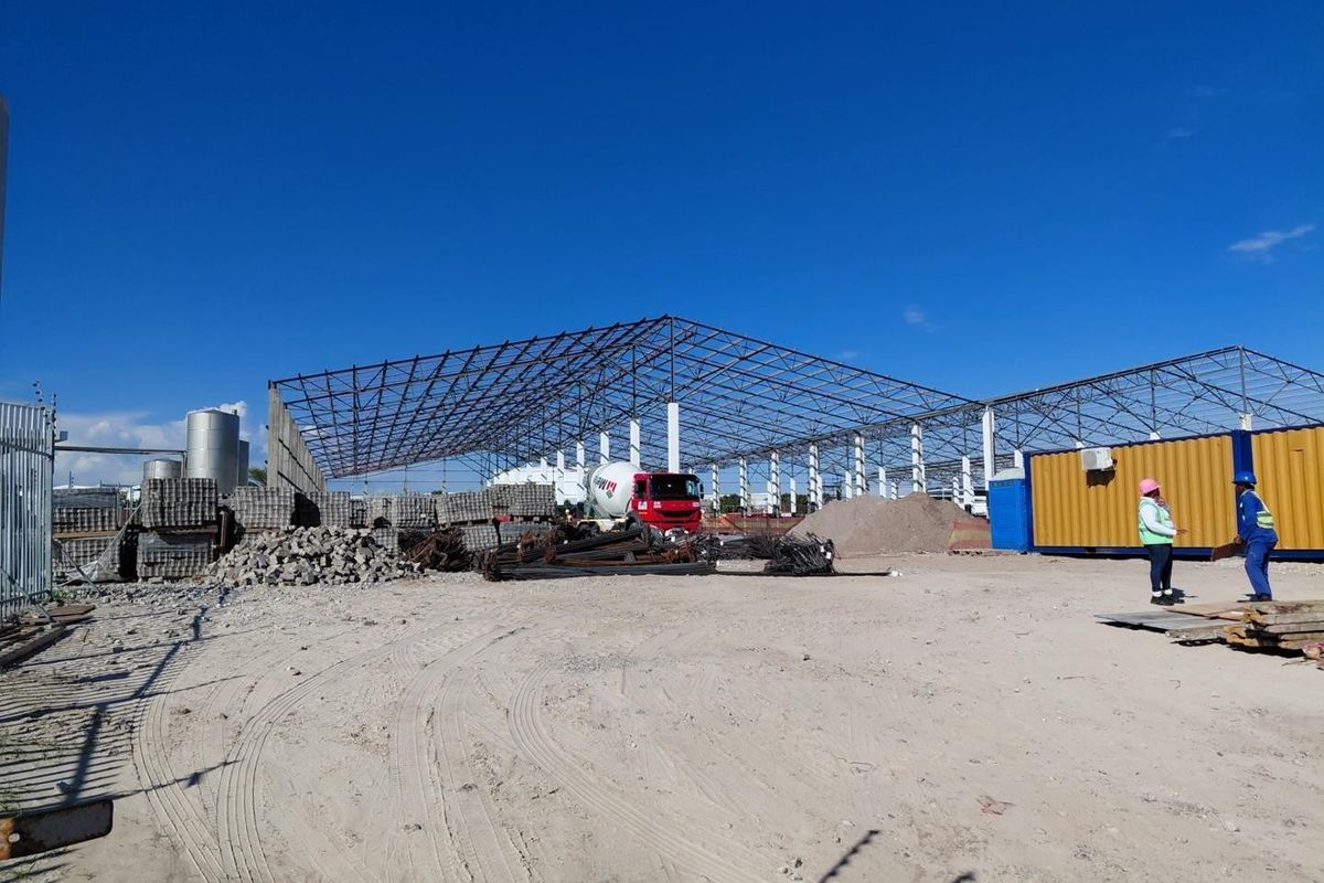 An industrial construction site featuring an expansive steel truss framework, storage containers, and raw materials under development.