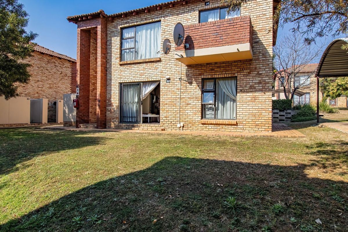 Rear view of a two-story face brick apartment building featuring a balcony, sliding door access, and an adjacent carport in a communal garden setting.