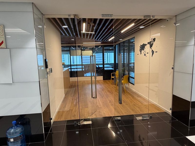 Modern office entrance featuring frameless glass double doors, wood-look flooring, and a contemporary slatted ceiling design.