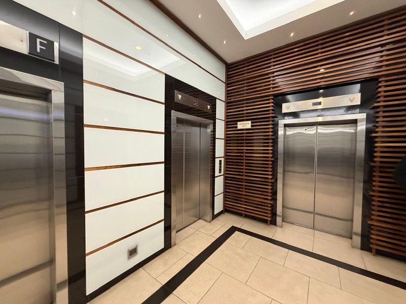 Modern office building elevator lobby featuring stainless steel lift doors, decorative timber slat wall paneling, and high-gloss white wall cladding.