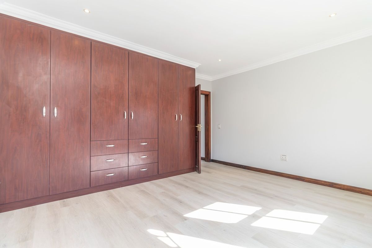 A bright, empty bedroom featuring extensive dark wood built-in cupboards, light-toned laminate flooring, and recessed ceiling lighting.