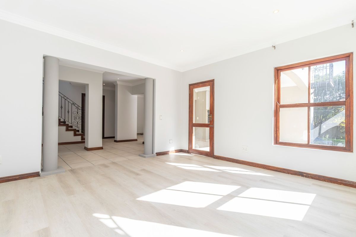 A bright, open-plan living area featuring light laminate flooring, structural columns, and a view toward the staircase with a decorative wrought iron balustrade.