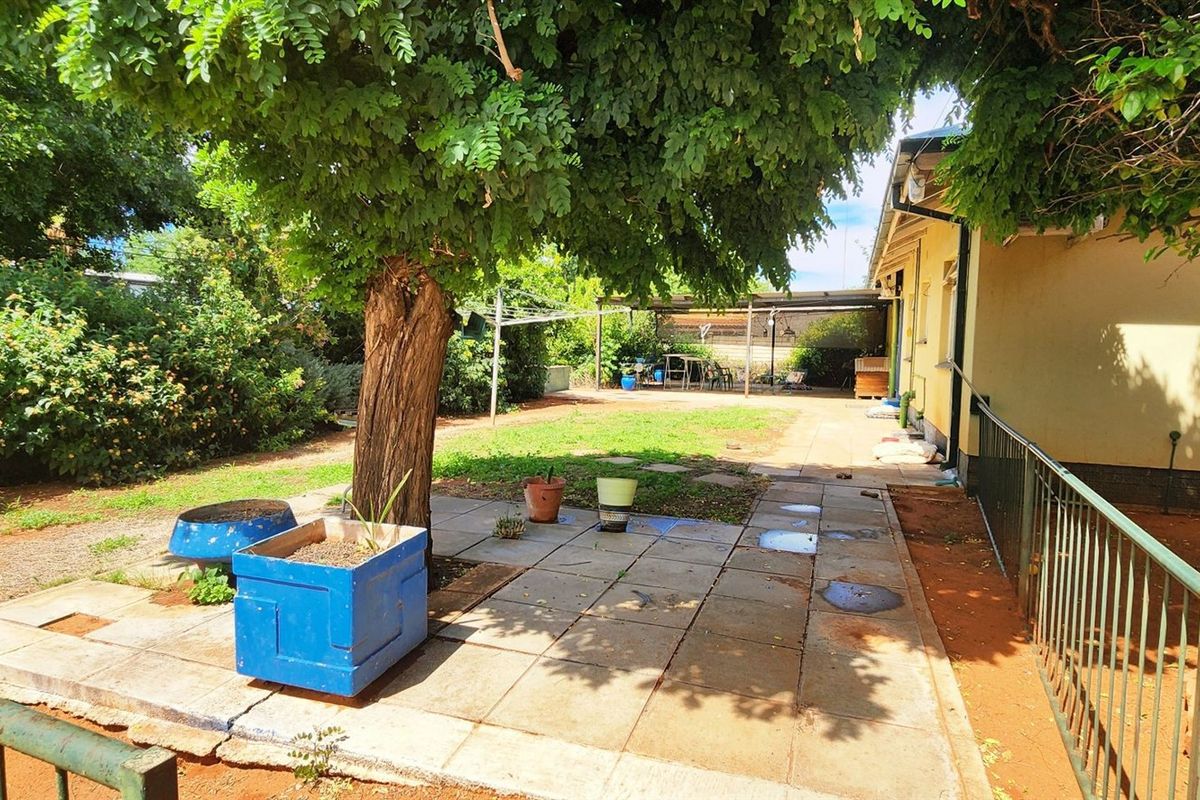 A rear garden area featuring a paved patio, mature tree, and metal security fencing along the side of the house.
