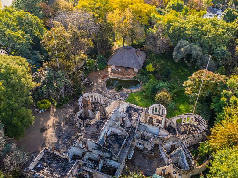 Aerial view of a residential property featuring a severely fire-damaged main structure, a swimming pool, and a separate thatched-roof outbuilding set within mature trees.