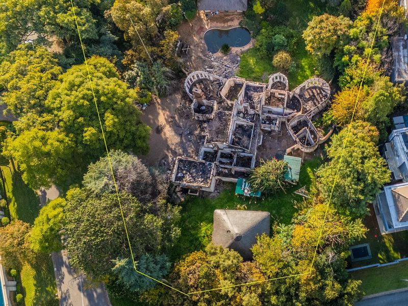 Aerial view of a residential property featuring a fire-damaged structure, mature trees, and a swimming pool on a large, wooded plot.