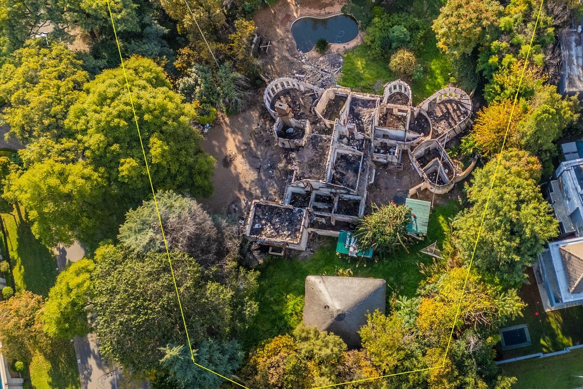 Aerial view of a residential property featuring a fire-damaged structure, mature trees, and a swimming pool on a large, wooded plot.