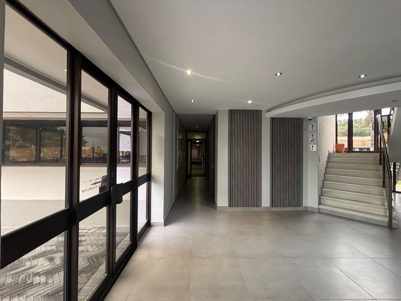 Modern office building entrance lobby featuring large glass doors, tiled flooring, decorative slatted wall paneling, and a staircase leading to upper levels.