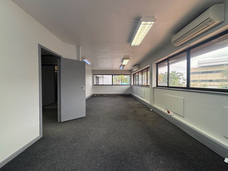 A vacant, open-plan office space featuring carpeted flooring, large windows for natural light, and wall-mounted air conditioning units.