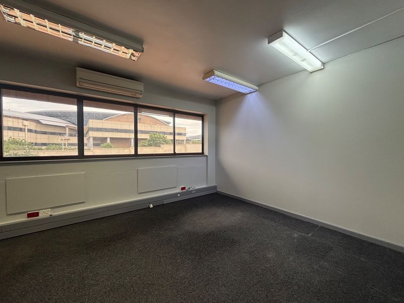 A clean, empty office space featuring carpet flooring, split-system air conditioning, and integrated fluorescent lighting with large windows providing natural light.
