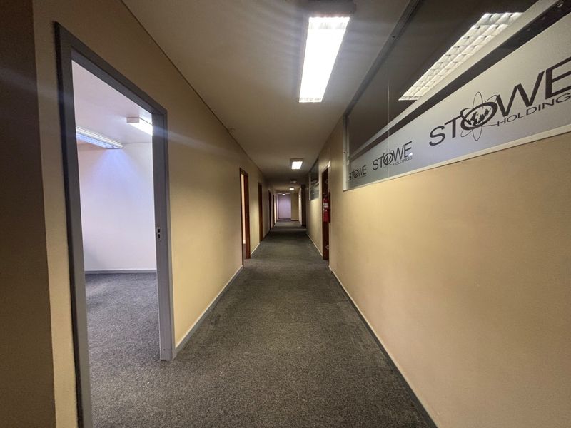 A long office hallway featuring carpeted flooring, fluorescent lighting, and glass partitions with corporate branding.