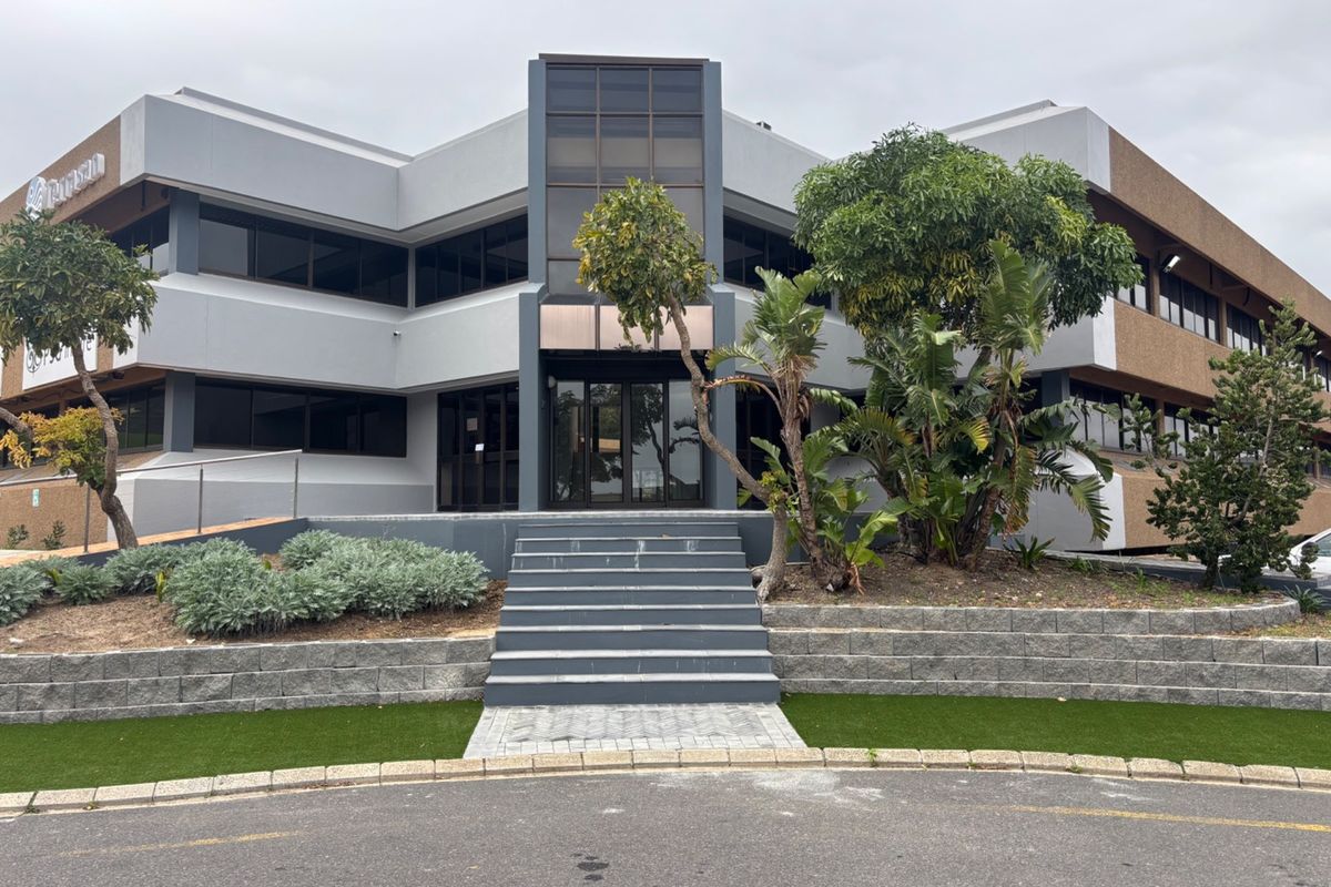 Front elevation of a modern multi-story office building featuring a central glass entrance, concrete retaining walls, and landscaped garden areas.