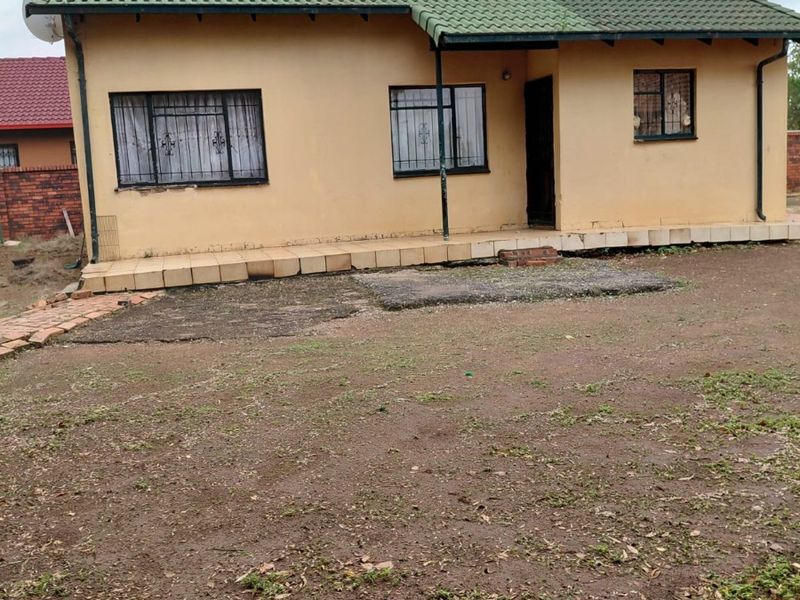 Front view of a single-story house featuring a tiled roof, solar water heater, and security bars on windows, set on a large unpaved yard.