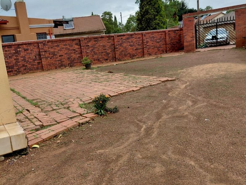 Front exterior view featuring a brick boundary wall, metal entrance gate, and a paved driveway area with an unmaintained dirt yard.