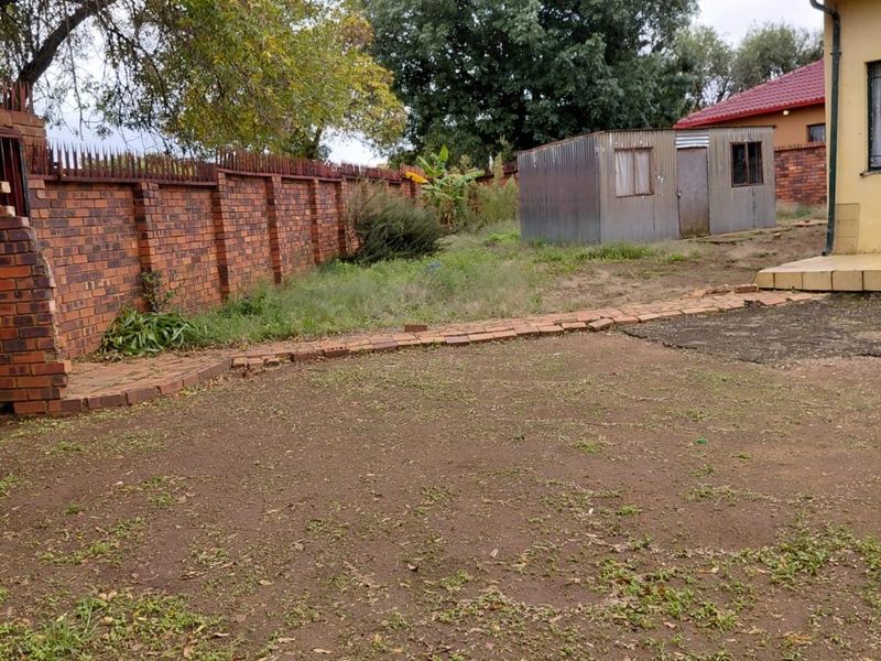 A spacious rear garden area featuring a brick boundary wall, a metal storage shed, and a paved walkway leading to the main house structure.
