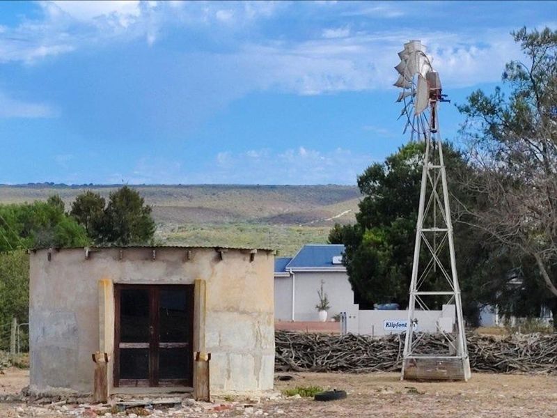A rural farm scene featuring a traditional metal windmill and a small, weathered circular outbuilding structure set against a landscape backdrop.