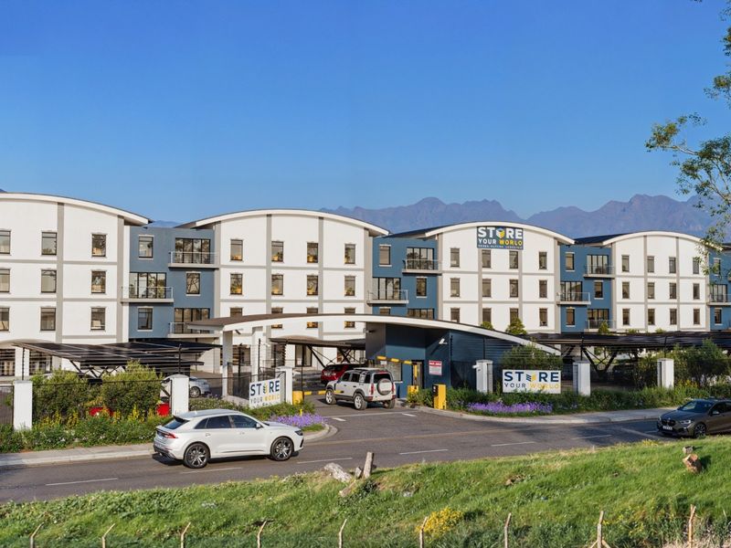 A wide-angle exterior view of a modern commercial storage facility complex featuring multi-story buildings, secure gated entrance, and covered parking areas.