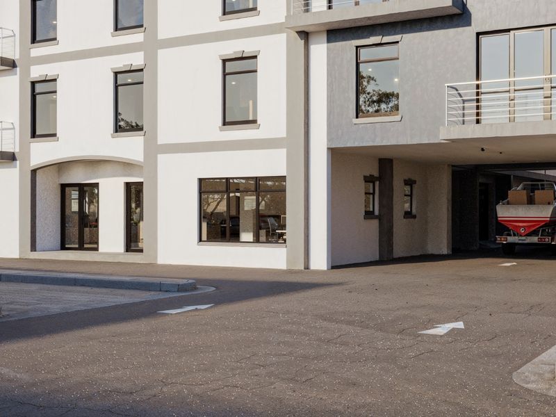 Modern commercial building exterior featuring a paved driveway, covered parking area, and large glass windows with a clean, contemporary facade.