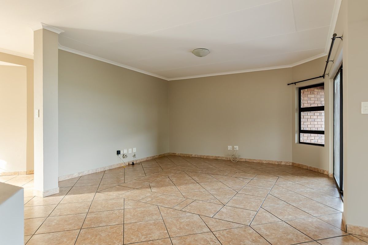 A spacious, empty living area featuring neutral wall tones, tiled flooring, and large windows providing natural light.