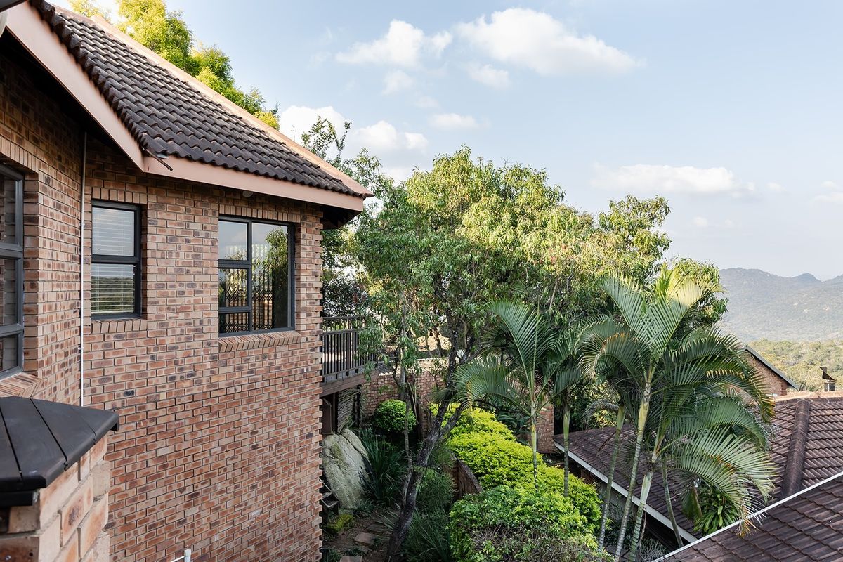 An elevated view of a brick-clad residential property featuring a tiled roof, wooden balcony, and lush surrounding garden with scenic mountain views.